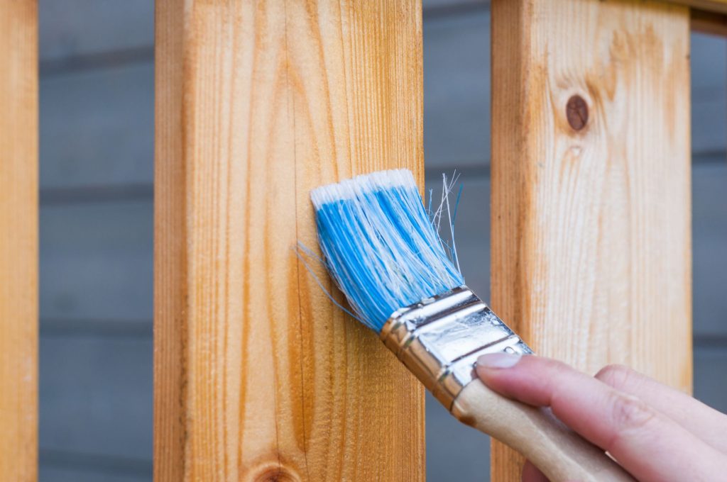 Main appliquant au pinceau une protection sur une planche de bois clair d'une balustrade extérieure