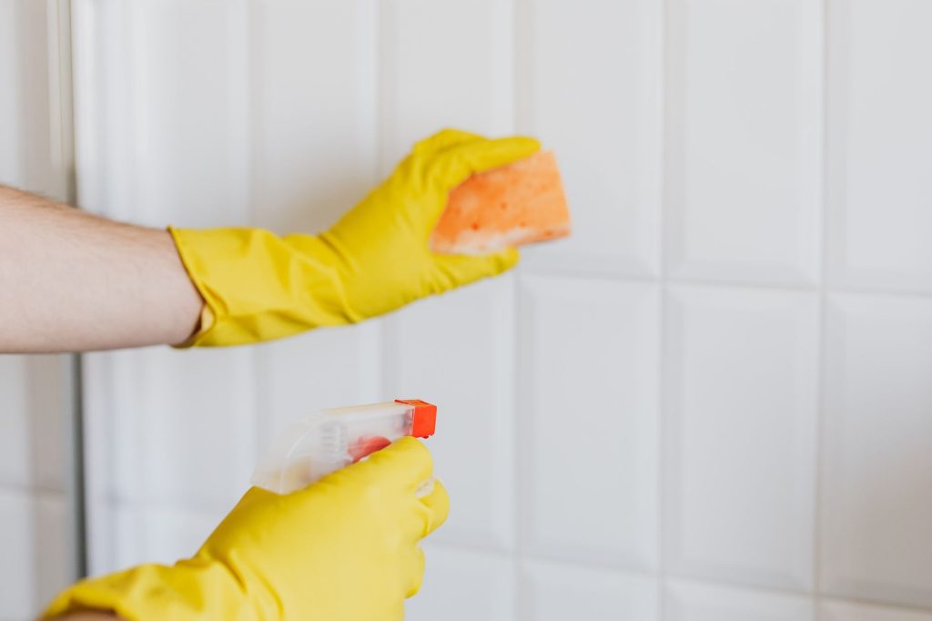 Mains gantées de jaune nettoyant un carrelage mural blanc avec une éponge et un pulvérisateur