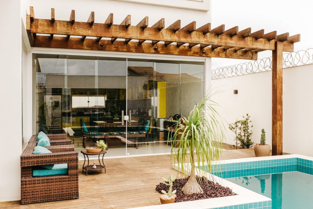 Pergola en bois adossée à une maison au-dessus d'une terrasse avec salon de jardin en bordure de piscine