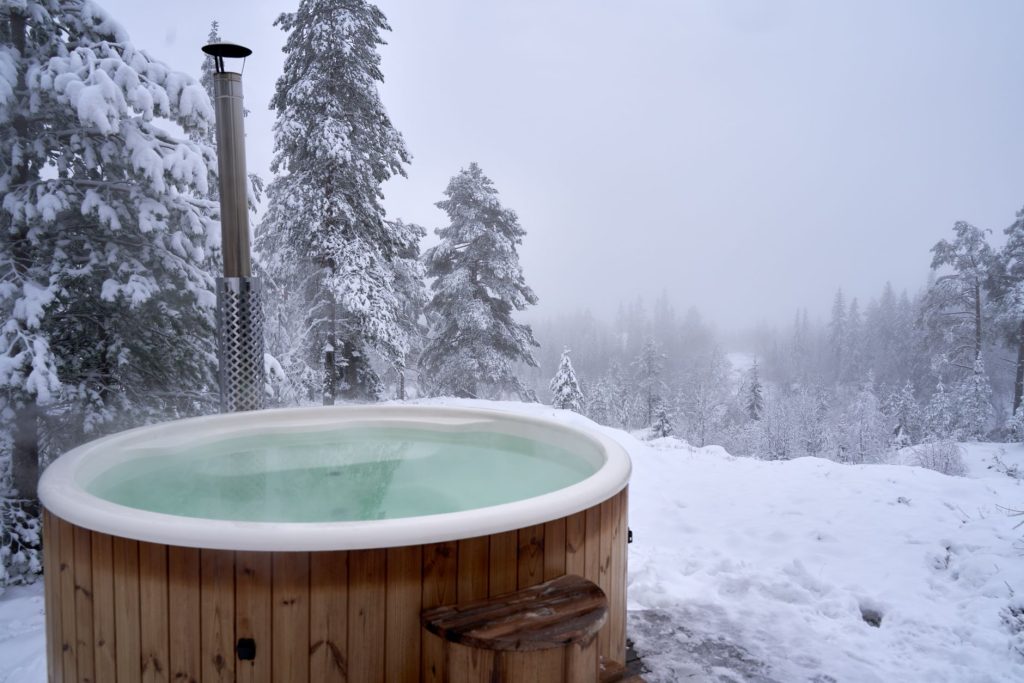 Spa en bois rempli d'eau chaude installé dans un jardin enneigé entouré de sapins