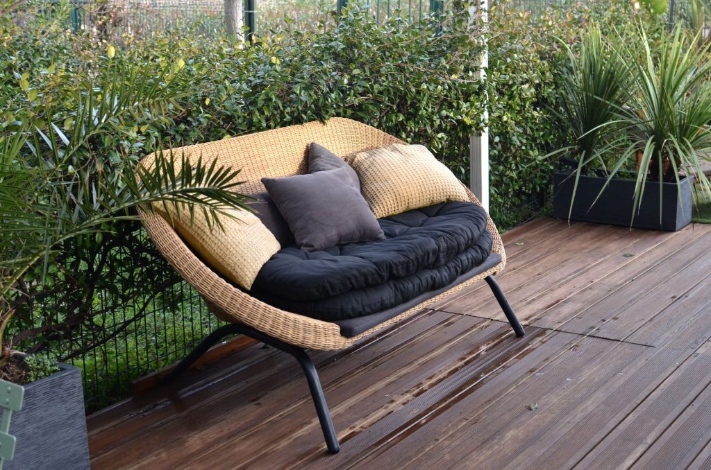Canapé en résine tressée avec coussins posé sur une terrasse en bois brun entourée de plantes