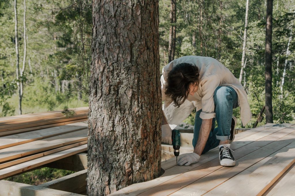 Homme vissant à la visseuse les lames d'une terrasse en bois clair autour d'un arbre